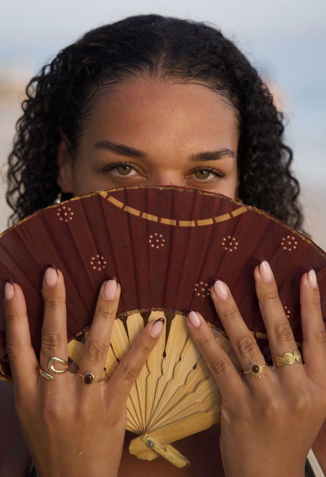 Woman on the beach holding a fan, wearing gold rings